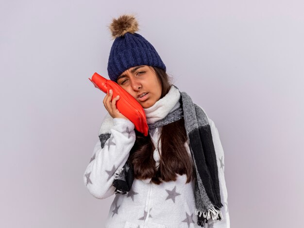 Triste joven enferma vistiendo gorro de invierno con bufanda poniendo bolsa de agua caliente en la mejilla aislado sobre fondo blanco.