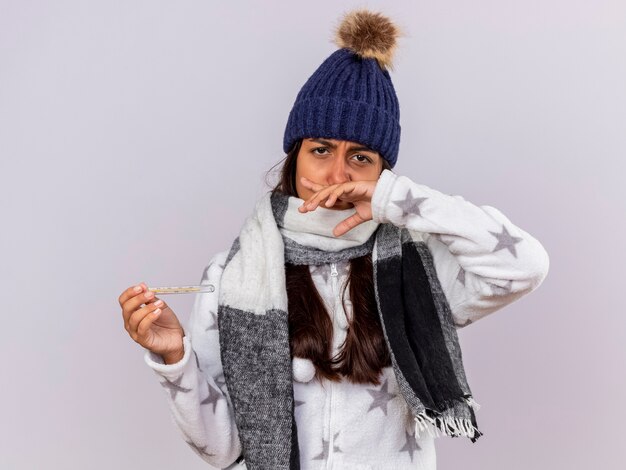 Triste joven enferma vistiendo gorro de invierno con bufanda sosteniendo el termómetro y limpiando la nariz con la mano aislado sobre fondo blanco.