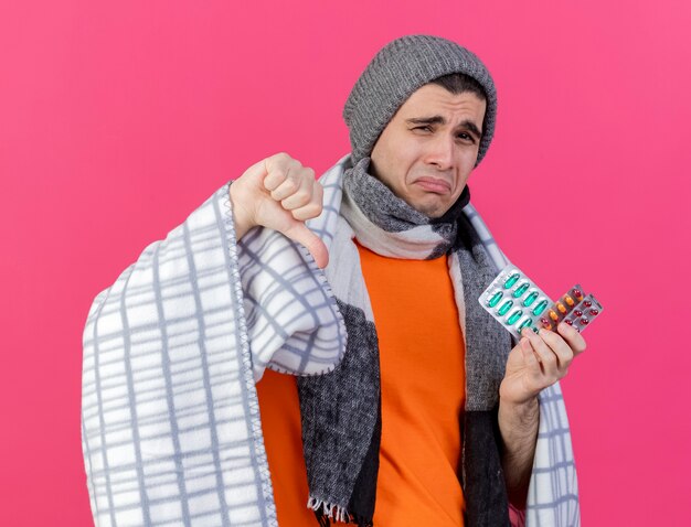 Triste joven enfermo con sombrero de invierno con bufanda envuelta en cuadros sosteniendo píldoras y mostrando el pulgar hacia abajo aislado en rosa