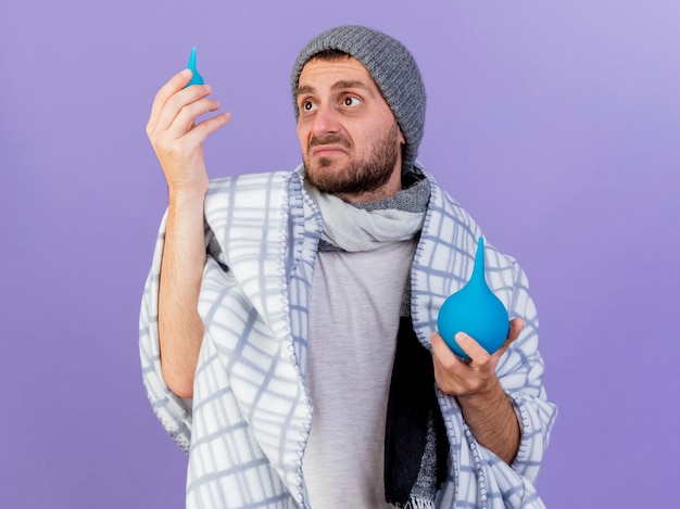 Triste joven enfermo con sombrero de invierno con bufanda levantando y mirando enema aislado sobre fondo púrpura