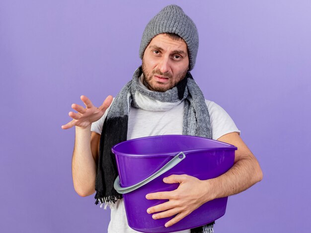 Triste joven enfermo con sombrero de invierno con bufanda sosteniendo un cubo de plástico aislado en púrpura