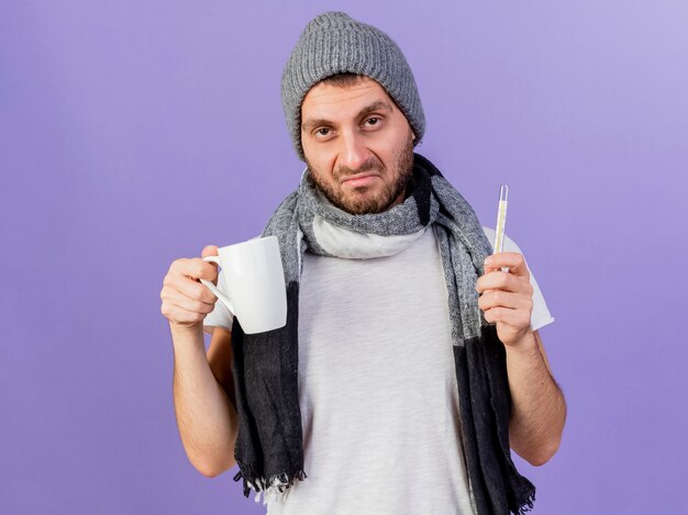 Triste joven enfermo con sombrero de invierno con bufanda sosteniendo termómetro con taza de té aislado en púrpura