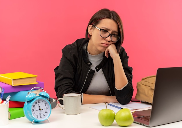 Triste joven estudiante con gafas sentado en el escritorio haciendo los deberes poniendo la mano en la cara mirando portátil aislado en la pared rosa