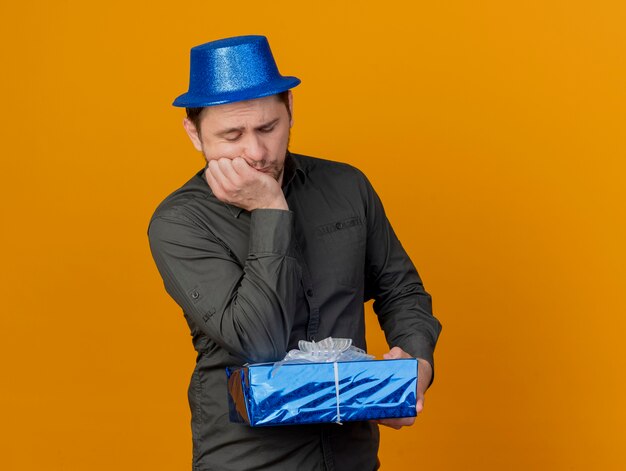 Triste joven fiestero con sombrero azul sosteniendo y poniendo el codo en la caja de regalo aislada en naranja