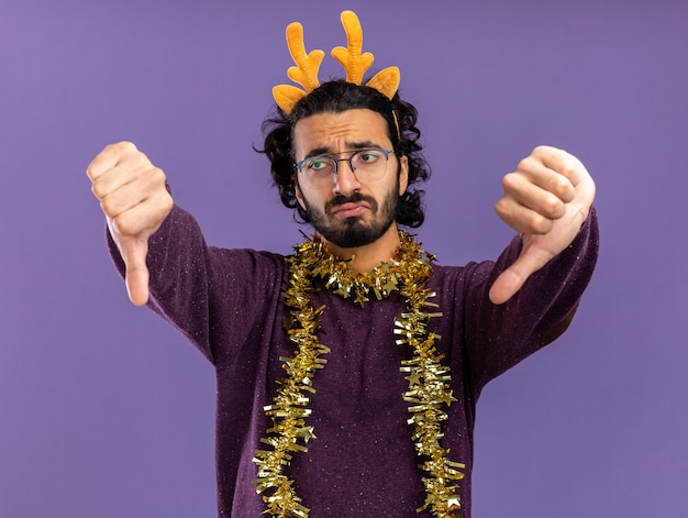 Triste joven guapo con aro de pelo de Navidad con guirnalda en el cuello mostrando los pulgares hacia abajo aislado sobre fondo azul.