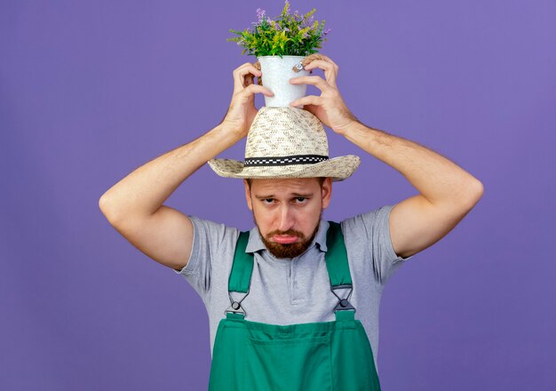 Triste joven guapo jardinero eslavo en uniforme y sombrero sosteniendo maceta en la cabeza aislada en la pared púrpura