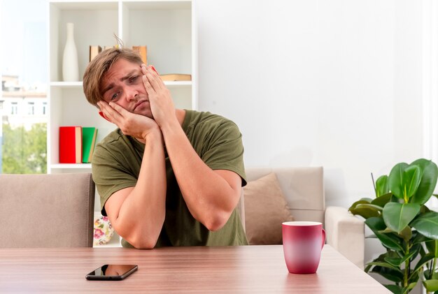 Triste joven guapo rubio se sienta a la mesa con taza y teléfono poniendo las manos en la cara mirando