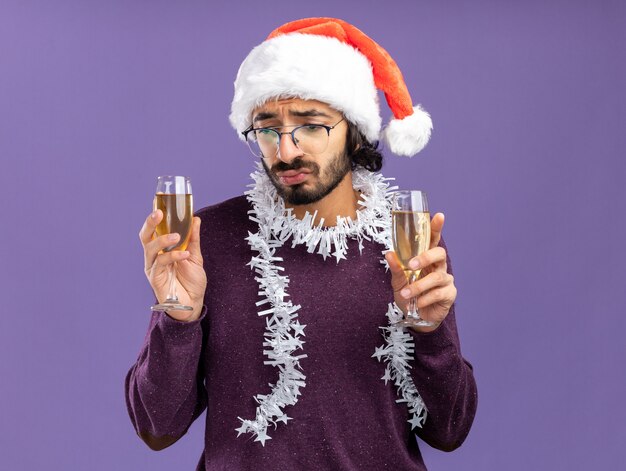 Triste joven guapo con sombrero de navidad con guirnalda en el cuello sosteniendo y mirando una copa de champán aislado sobre fondo azul.