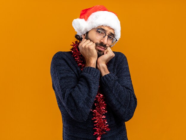 Triste joven guapo con sombrero de navidad con guirnalda en el cuello tomados de la mano en las mejillas aisladas sobre fondo naranja con espacio de copia