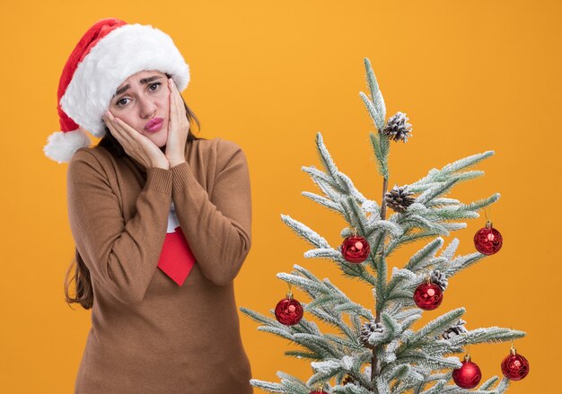 Triste joven hermosa niña con sombrero de navidad con corbata de pie cerca del árbol de navidad poniendo las manos en las mejillas aisladas sobre fondo naranja