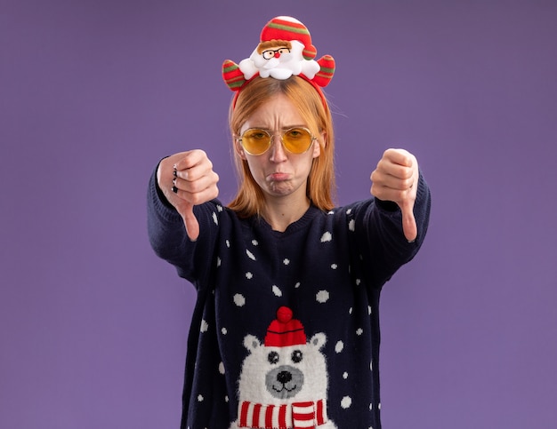 Triste joven hermosa niña vistiendo un suéter de Navidad y un aro de pelo de Navidad con gafas mostrando los pulgares hacia abajo aislado sobre fondo púrpura