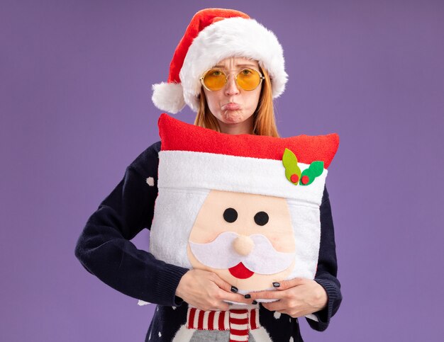 Triste joven hermosa niña vistiendo suéter de Navidad y sombrero con gafas sosteniendo almohada de Navidad aislado en la pared púrpura