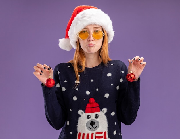 Triste joven hermosa niña vistiendo un suéter de Navidad y sombrero con gafas sosteniendo bolas de Navidad aisladas sobre fondo púrpura