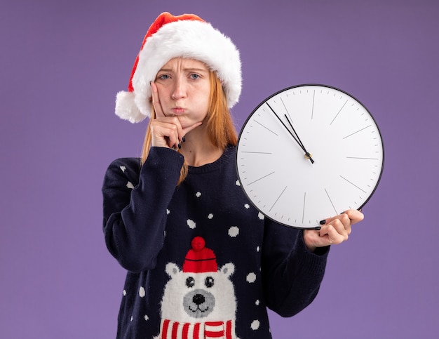 Triste joven hermosa niña vistiendo un suéter de Navidad y un sombrero sosteniendo el reloj de pared poniendo la mano en la mejilla aislado sobre fondo púrpura