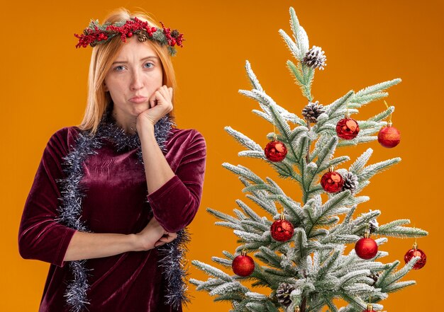 Triste joven hermosa de pie cerca del árbol de Navidad con vestido rojo y corona con guirnalda en el cuello poniendo la mano en el mentón aislado sobre fondo naranja