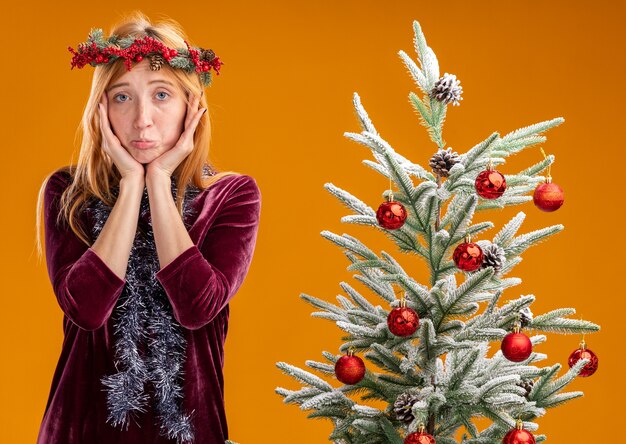 Triste joven hermosa de pie cerca del árbol de Navidad con vestido rojo y corona con guirnalda en el cuello poniendo las manos alrededor de la cara aislada sobre fondo naranja