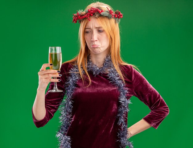 Triste joven hermosa vestida de rojo con corona y guirnalda en el cuello sosteniendo y mirando una copa de champán aislado sobre fondo verde