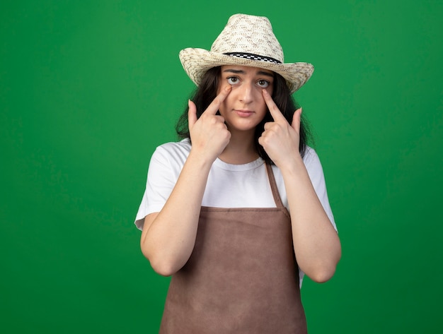 Triste joven jardinero mujer morena con gafas ópticas y uniforme con sombrero de jardinería tira hacia abajo los párpados aislados en la pared verde