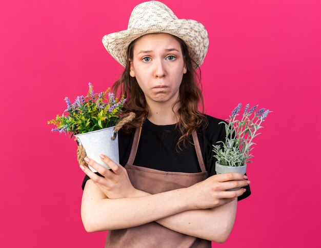 Triste joven jardinero con sombrero de jardinería sosteniendo y cruzando flores en macetas