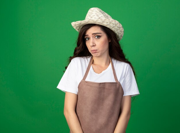 Triste joven jardinero en uniforme con sombrero de jardinería mira al frente aislado en la pared verde