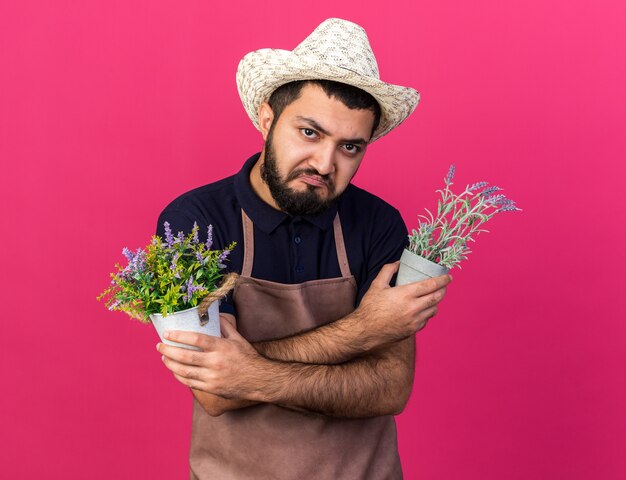 Triste joven jardinero varón caucásico vistiendo sombrero de jardinería sosteniendo macetas cruzando los brazos aislado en la pared rosa con espacio de copia