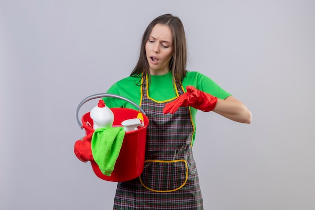 Triste joven de limpieza con uniforme en guantes rojos apunta a herramientas de limpieza en su mano sobre fondo blanco aislado