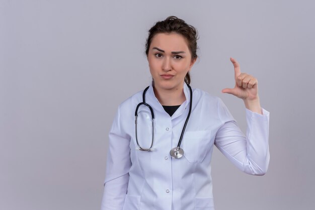 Triste joven médico con bata médica con estetoscopio muestra el tamaño en la pared blanca