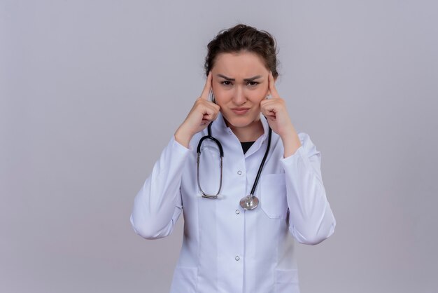 Triste joven médico con bata médica con estetoscopio puso sus dedos en la frente en la pared blanca