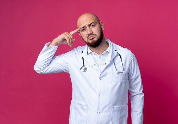 Triste joven médico calvo vistiendo bata médica y estetoscopio poniendo el dedo en la frente aislado en rosa