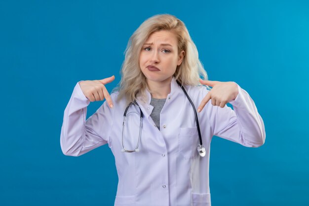 Triste joven médico con estetoscopio en bata médica apunta a sí misma en la pared azul