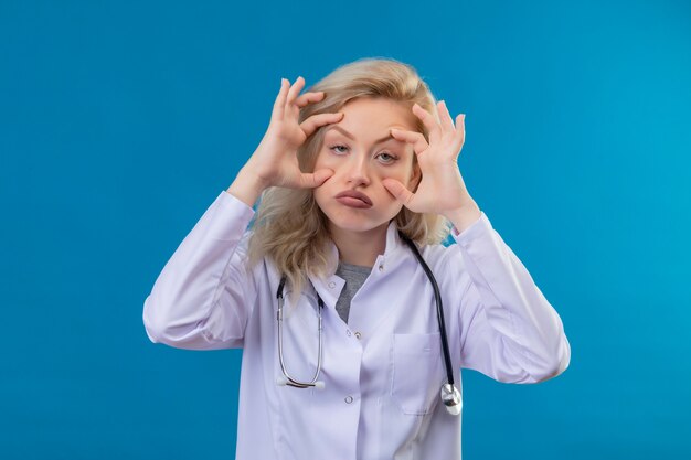 Triste joven médico con estetoscopio en bata médica poniendo las manos alrededor de los ojos en la pared azul