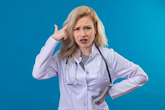 Triste joven médico con estetoscopio en bata médica puso su dedo en la cabeza en la pared azul