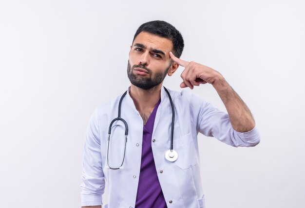 Triste joven médico con estetoscopio bata médica puso su dedo en la frente sobre fondo blanco aislado