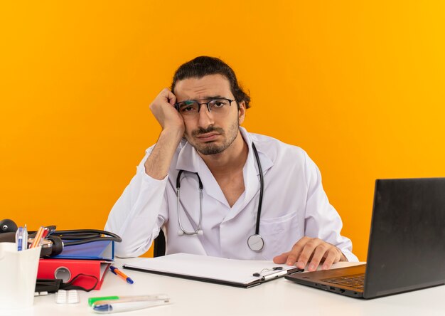 Triste joven médico con gafas médicas vistiendo bata médica con estetoscopio sentado