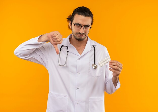 Triste joven médico con gafas médicas vistiendo bata médica con estetoscopio sosteniendo pastillas con el pulgar hacia abajo en la pared amarilla aislada con espacio de copia