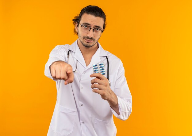 Triste joven médico con gafas médicas vistiendo bata médica con estetoscopio sosteniendo píldoras y mostrando gesto