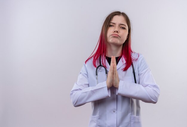 Triste joven médico vestida con un estetoscopio bata médica mostrando gesto de oración sobre fondo blanco aislado con espacio de copia