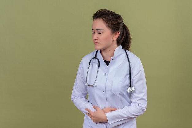 Triste joven médico vistiendo bata médica con estetoscopio agarrando el estómago en la pared verde