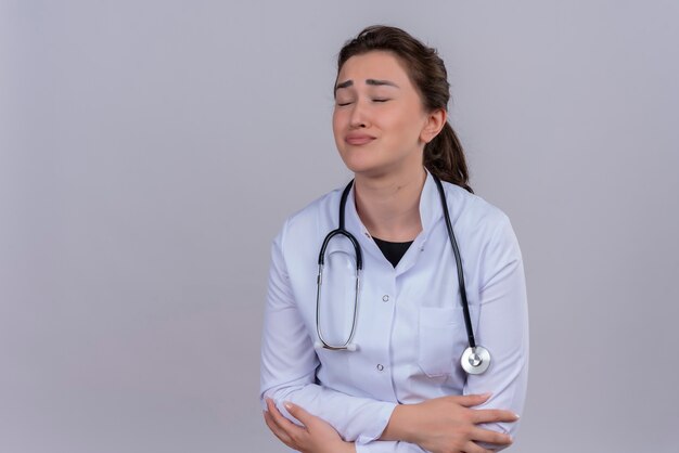 Triste joven médico vistiendo bata médica con estetoscopio cruzando las manos sobre la pared blanca