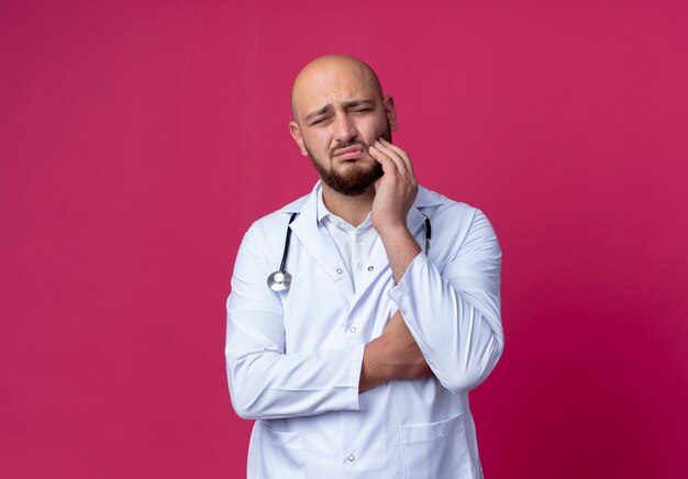 Triste joven médico vistiendo bata médica y estetoscopio poniendo la mano sobre el dolor de muelas aislado en rosa