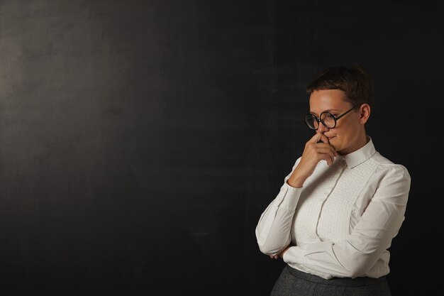 Triste joven profesora con blusa blanca y falda gris sumida en sus pensamientos junto a una pizarra en blanco