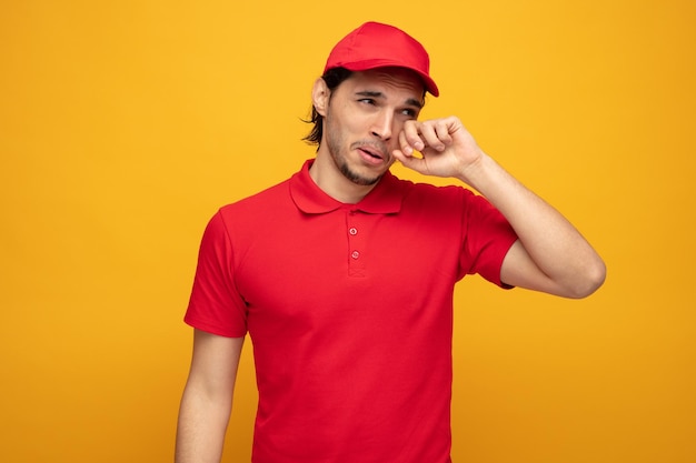 Triste joven repartidor con uniforme y gorra mirando a un lado llorando limpiándose los ojos con la mano aislada en el fondo amarillo