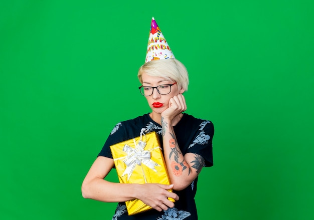 Triste joven rubia fiestera con gafas y gorro de cumpleaños con caja de regalo mirando hacia abajo aislado sobre fondo verde con espacio de copia