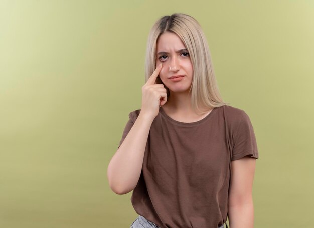 Triste joven rubia gesticulando llorar con el dedo en el ojo en el espacio verde aislado con espacio de copia