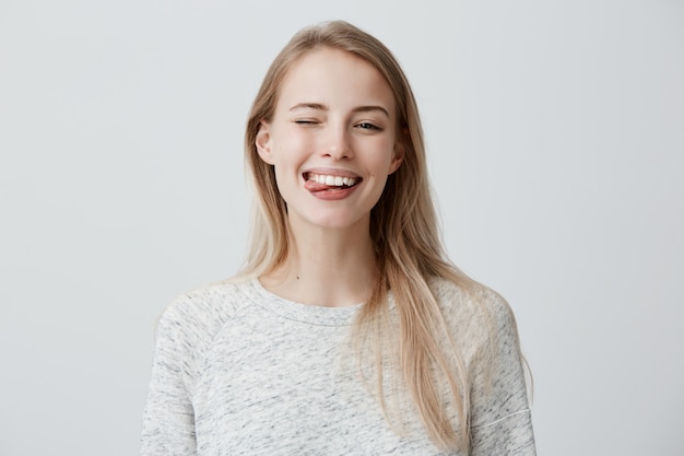 Triste joven rubia con el pelo teñido, vestida casualmente haciendo muecas, parpadeando y sacando la lengua. mujer positiva divirtiéndose en el interior