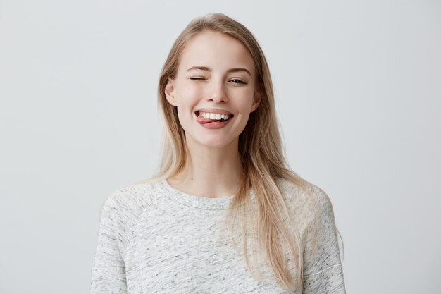 Triste joven rubia con el pelo teñido, vestida casualmente haciendo muecas, parpadeando y sacando la lengua. Mujer positiva divirtiéndose en el interior