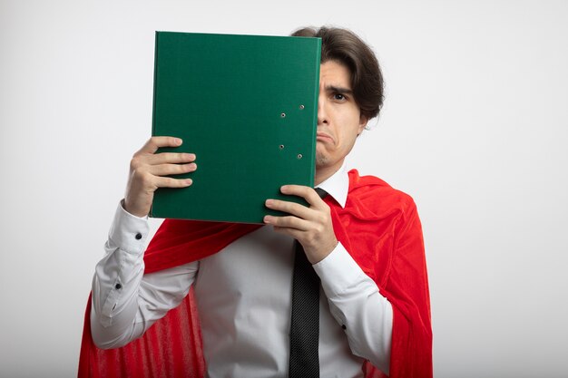 Triste joven superhéroe con corbata cubrió la cara con portapapeles aislado sobre fondo blanco.