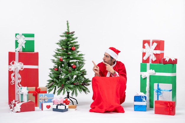 Triste joven vestido como Papá Noel con regalos y árbol de Navidad decorado apuntando algo en el lado derecho sobre fondo blanco.
