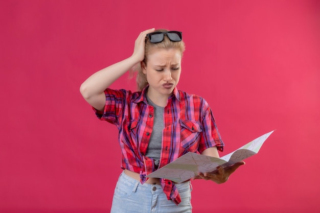 Foto gratuita triste joven viajera con camisa roja y gafas en la cabeza mirando al mapa puso su mano sobre la cabeza en la pared rosa aislada