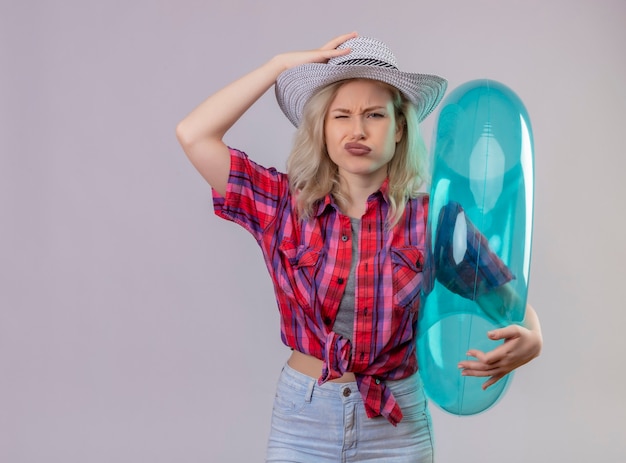 Triste joven viajera vestida con camisa roja con anillo inflable puso su mano en el sombrero en la pared blanca aislada